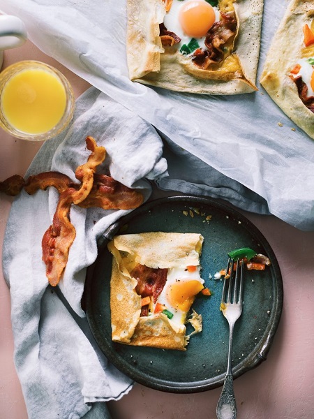 A Savory Crepe Pocket on a plate with a side of bacon and a glass of orange juice.