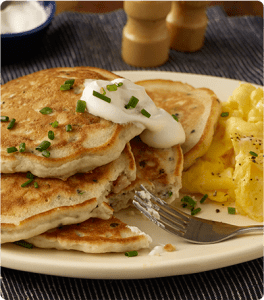 A stack of Black Forest and Seed Pancakes topped with sour cream and scallions and a side of scrambled eggs.