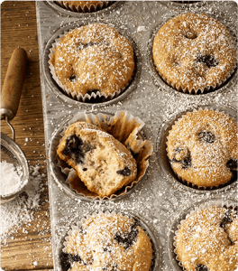 A muffin pan full of Blueberry Banana Muffins that are sprinkled with powdered sugar and one of the muffins has a bite taken out of it.