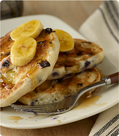 A stack of Blueberry Banana Pancakes topped with syrup and banana slices.