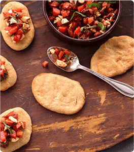 Bruschetta bites sit on a serving board next to to a bowl of fiery bruschetta toppings.