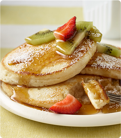 A stack of French Vanilla Pancakes topped with syrup, sliced strawberries and kiwi, and sprinkled with powdered sugar.