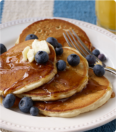 A stack of Gluten Free Classic Pancakes topped with butter, syrup and blueberries on a white plate with a silver fork.