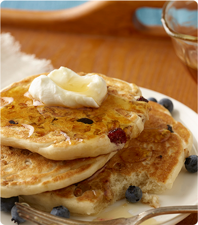 A stack of pancakes topped with granola, syrup, blueberries and butter.