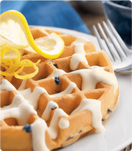 A Lemon Cream Blueberry Waffle, drizzled with cream and garnished with a slice of lemon, sits on a white plate.