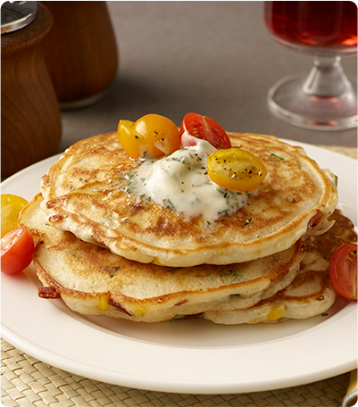 A stack of Roasted Corn and Basil Bacon Pancakes topped with butter, cherry tomatoes and chopped basil.