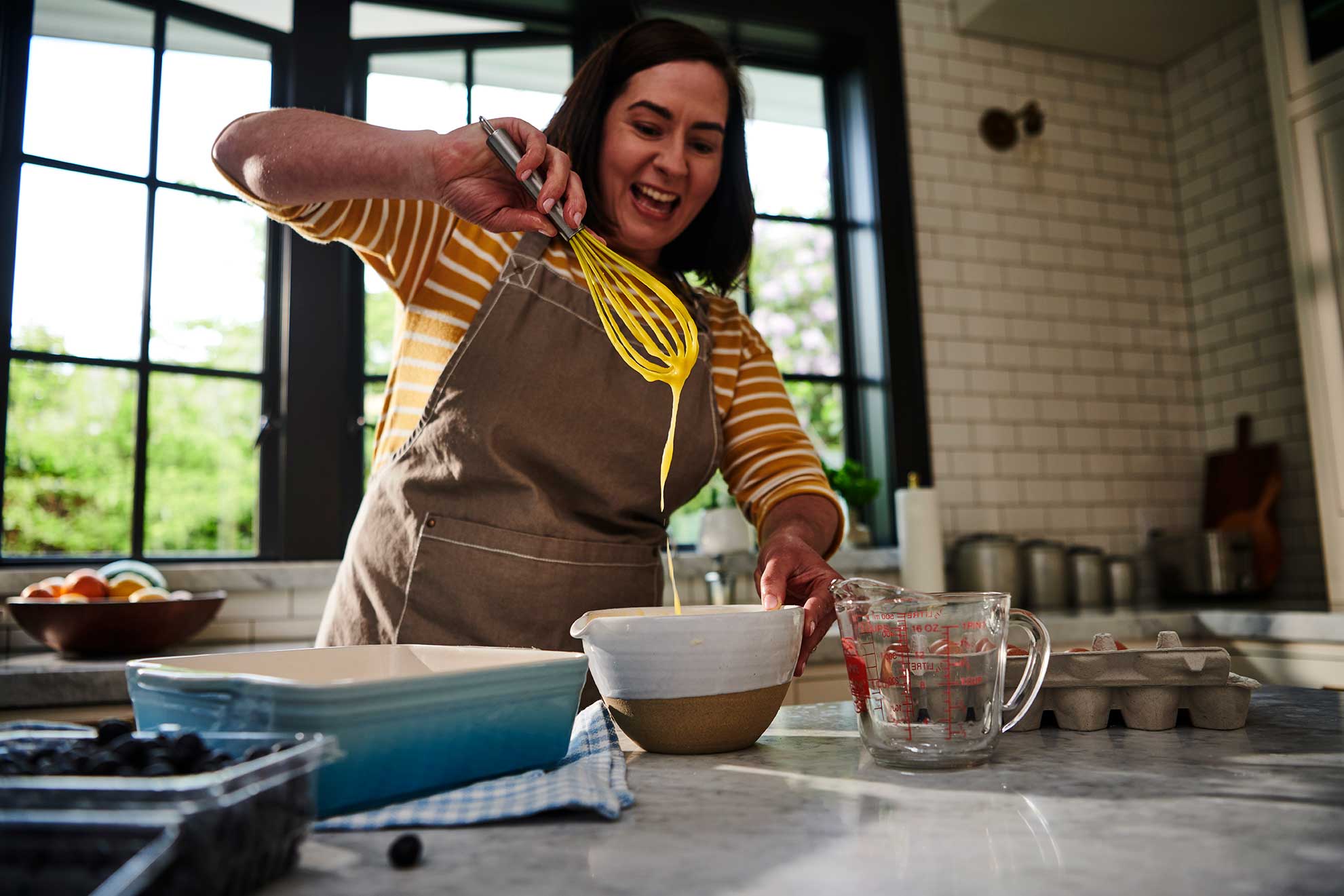 A woman in the kitchen smiling as she prepares Pumpkin Pie Bar batter with a whisk and mixing bowl.