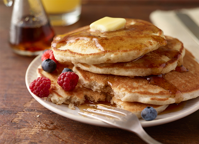 A white plate with a stack of gluten free buttermilk pancakes that's topped with butter, syrup, and fresh fruit.