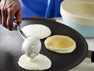 A person uses an ice cream scoop to place buttermilk pancake mix on a hot griddle.