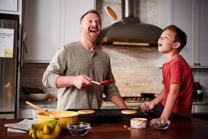 A dad joyously flips a buttermilk pancake while his kid watches with a smile.