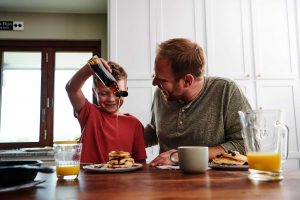 A smiling kid pours syrup on a stack of Krusteaz buttermilk pancakes while sitting at the table with his dad.