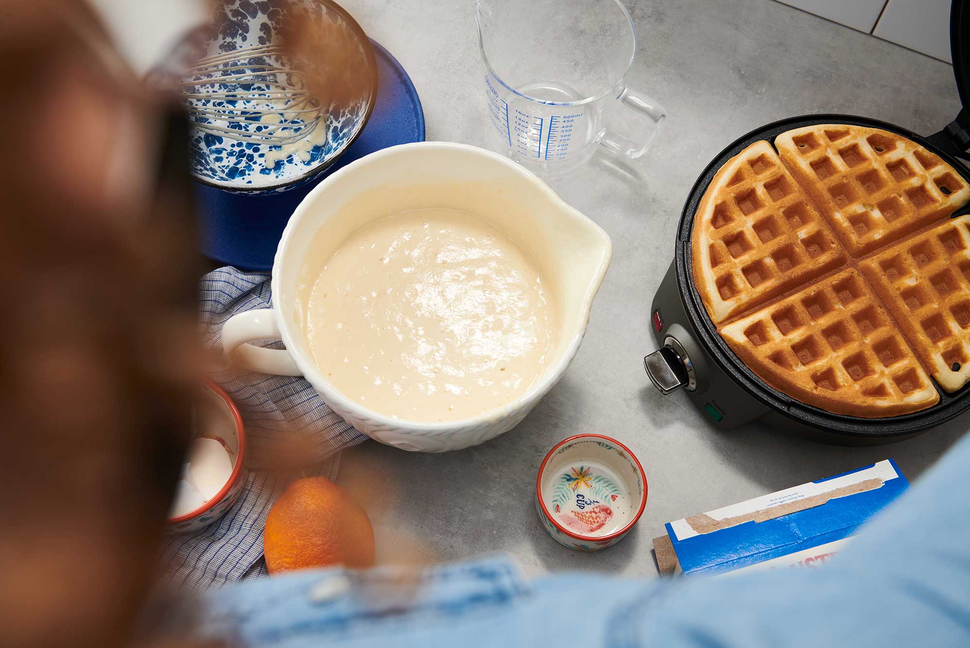 A white mixing bowl full of batter sits next to a waffle maker on a kitchen counter.