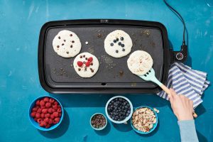 Four Krusteaz buttermilk pancakes cook on a griddle, each with different toppings, like chocolate chips, raspberries, blueberries, and almond slices.