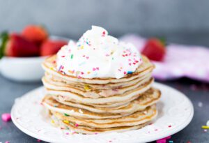 A stack of Coconut Mardi Gras Pancakes topped with whipped cream and sprinkles on a white plate.