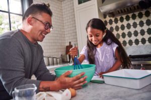 A young girl uses a blue whisk to mix batter while her father holds the green mixing bowl.