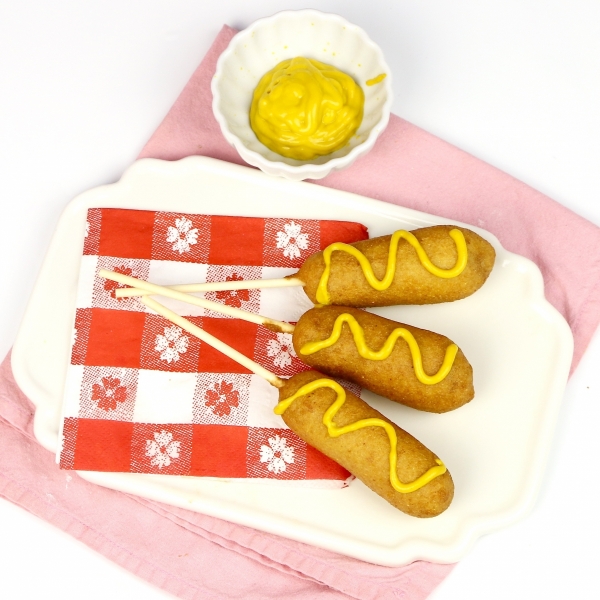 Three Mini Corn Dogs topped with mustard on a plate and served with a small bowl of mustard.