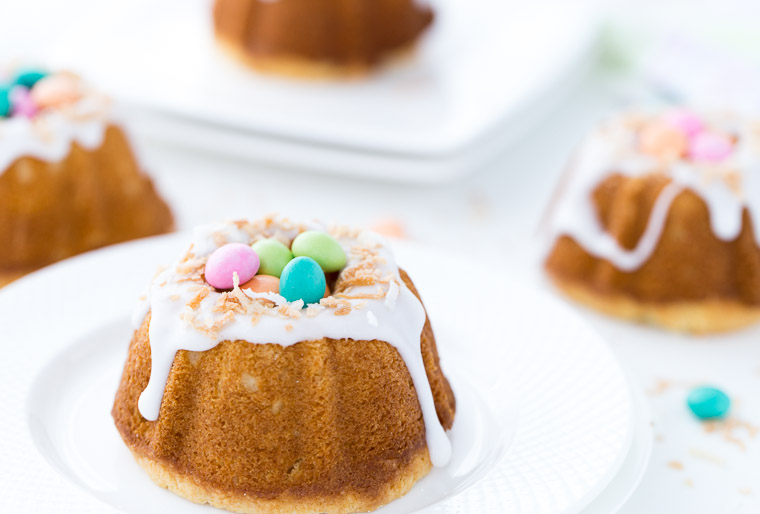 Several mini Coconut Lemon Bundt Cakes topped with a lemon glaze, coconut flakes and multicolored chocolate Easter eggs.