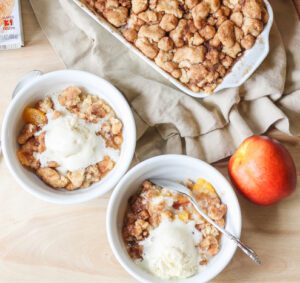 Two bowls of Peach Snickerdoodle Crumble topped with fresh peaches and vanilla ice cream.