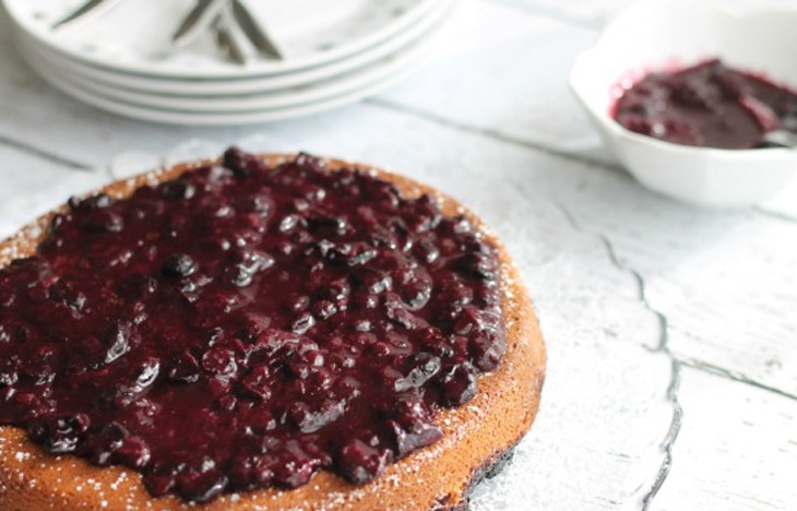 A Gluten Free Blueberry Coffee Cake with Blueberry Compote on a glass dish on a white, wooden table.