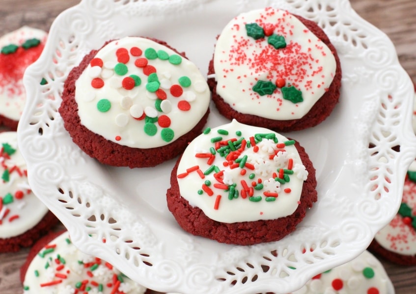 A plate of Red Velvet Christmas Cookies topped with a cream cheese frosting and festive sprinkles.