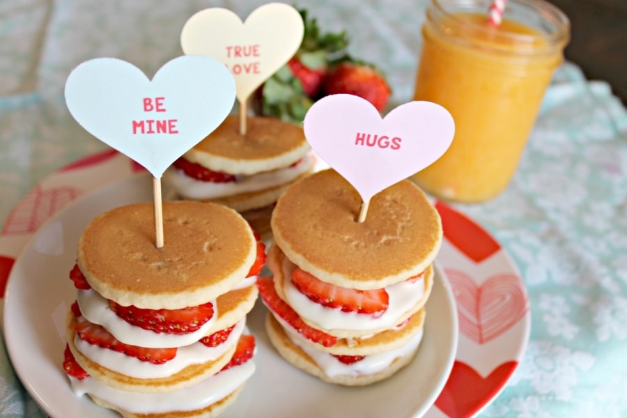 Three stacks of Strawberry Shortcake Mini Stacks paired with a glass of orange juice. In each stack of pancakes is a heart shaped decoration.