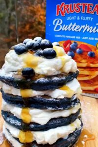 A stack of Midnight Blueberry Pancakes with whipped cream in between each pancake and topped with whipped cream, syrup and blueberries.