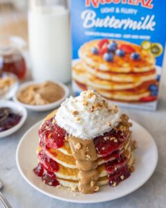 A stack of Peanut Butter and Jelly Pancakes topped with peanut butter, jelly, whipped cream and chopped peanuts.