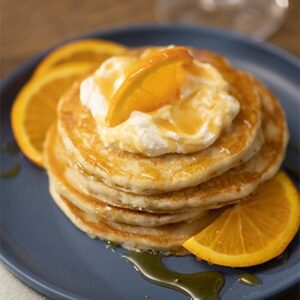 A stack of Mimosa Pancakes topped with whipped cream, orange slices and syrup.