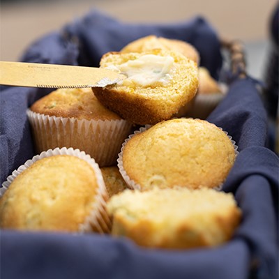 A basket of Elote Cornbread Muffins with one muffin being topped with butter.