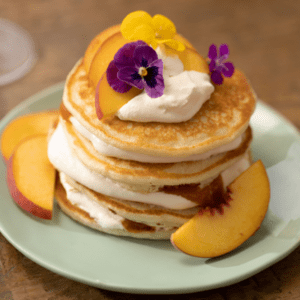 A stack of Peach Bellini Pancakes topped with whipped cream, peach slices and decorative flowers as garnish.