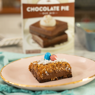 A chocolate coconut pie bar adorned with eastern candy sits on a white plate in front of a box of Krusteaz Chocolate Pie Bar mix.