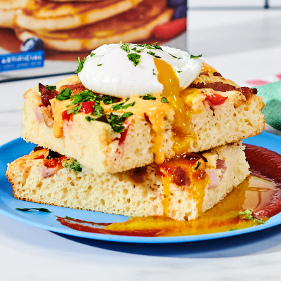 A plate of Savory Sheet Pan Pancakes topped with crumbled bacon, diced ham, cooked sausage, red bell peppers, green onions, a poached egg, queso and chopped cilantro.