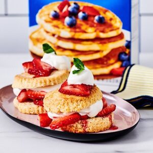 Two shortcakes layered with fluffy cream and strawberries sit on a plate next to a box of Krusteaz Buttermilk Pancake Mix.