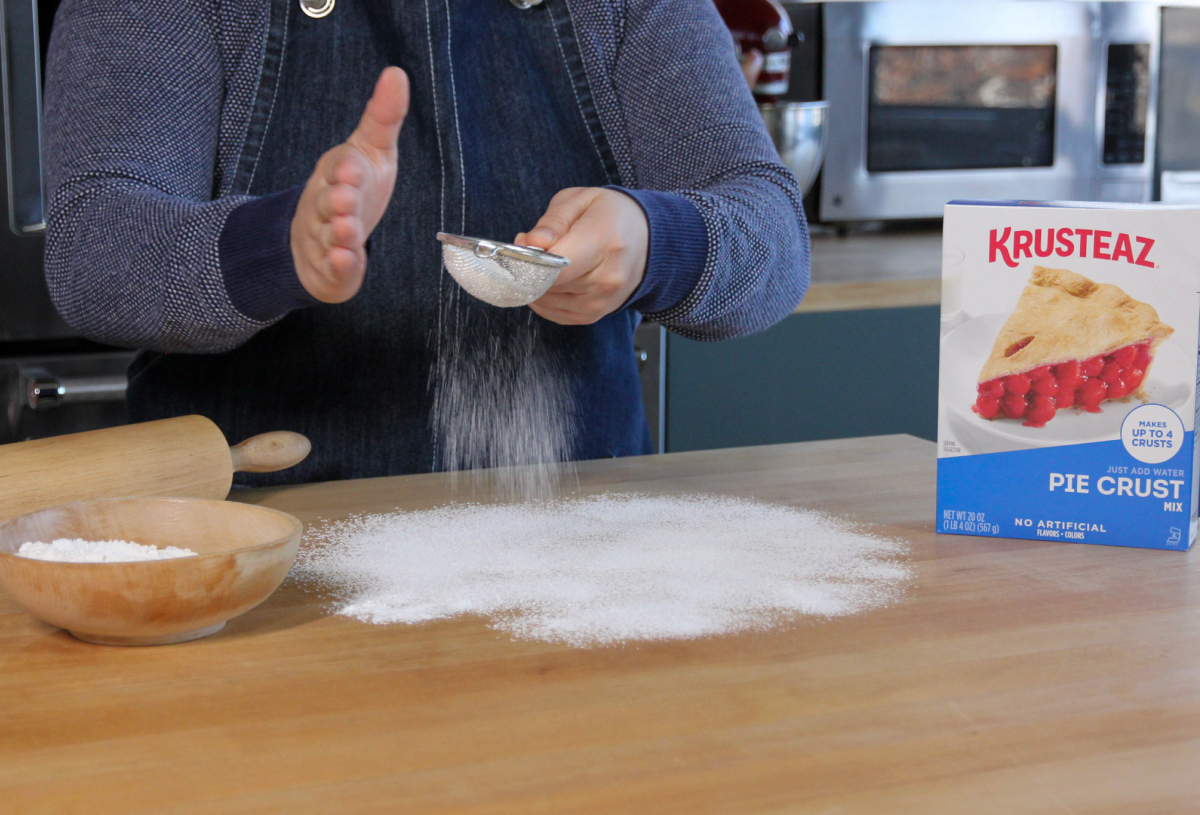 A cook sifting sugar onto a counter top with a box of Krusteaz Pie Crust next to them
